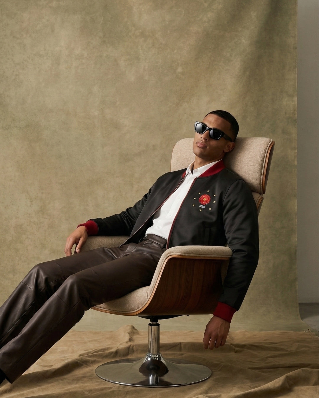 Man sitting in a chair wearing sunglasses and a dark jacket with a red emblem.