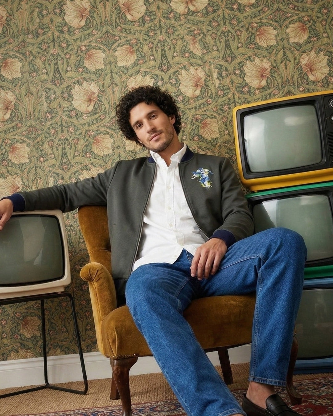 Man sitting on a vintage armchair with stacked old TVs against a floral wallpapered wall.