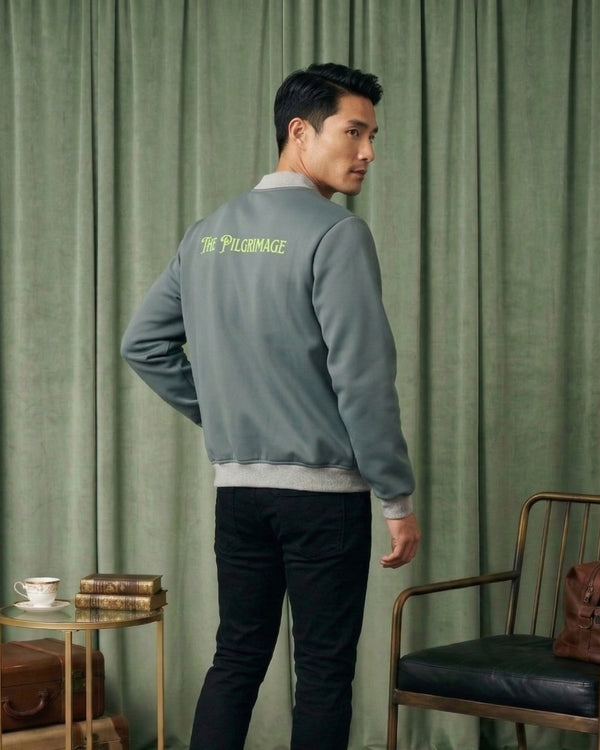 Man wearing a gray jacket with 'The Pilgrimage' logo, standing in a room with green curtains and a chair.