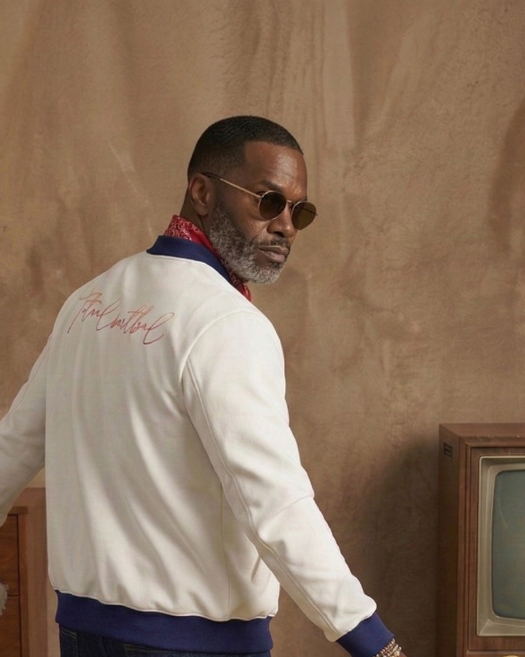 Man wearing a white jacket with text, sunglasses, and a red scarf against a beige wall.