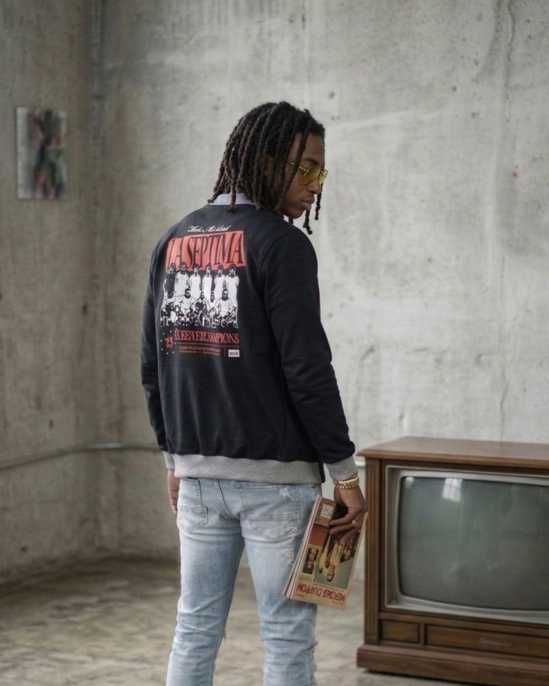 Person wearing a black jacket with red and white text, standing in a room with a vintage television and concrete wall.