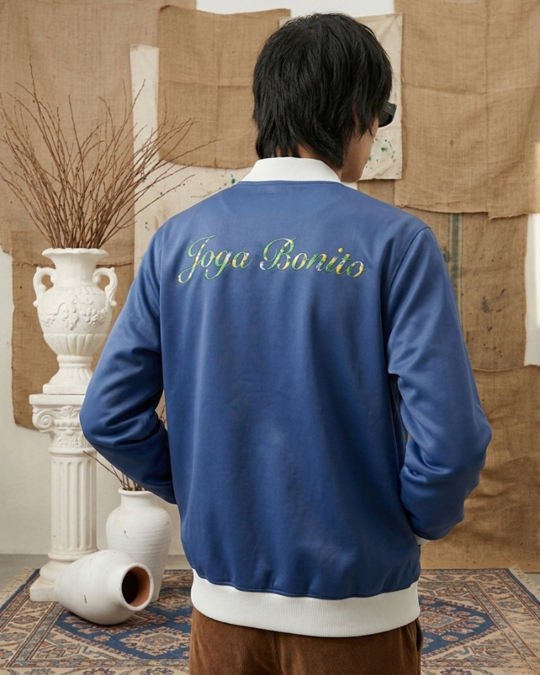 Person wearing a blue jacket with 'Joya Bonito' text, standing in a room with decorative elements.